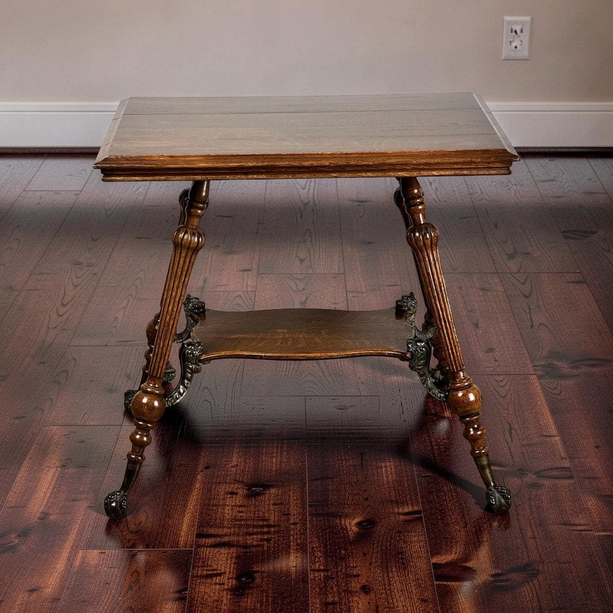 An Antique Victorian Oak Lamp Stand with Bronze Lions Head Mountings (c. Late 19th Century) stands on a dark hardwood floor beside a beige wall with white baseboard and outlet, featuring a lower shelf and ornate legs.
