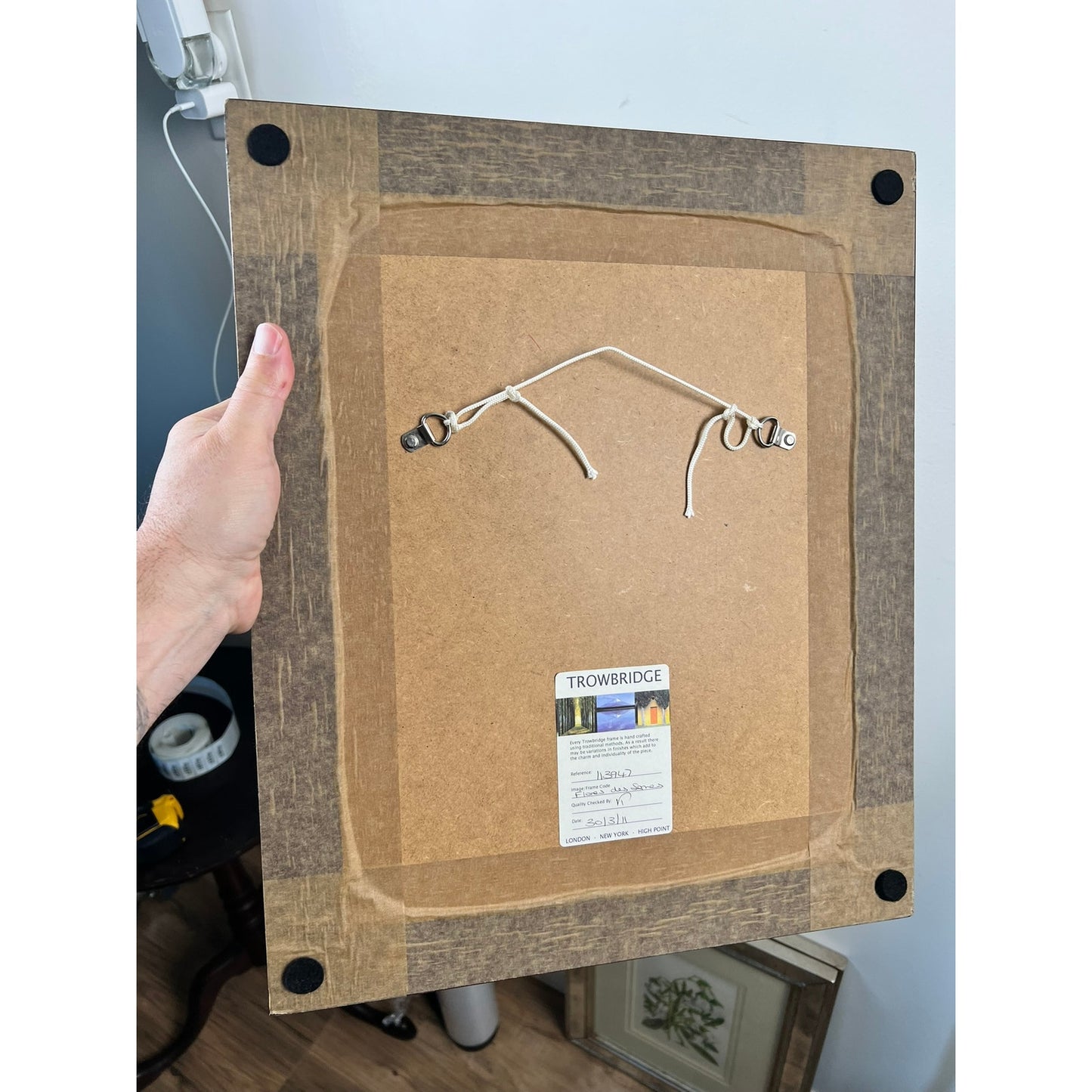A hand holds the back of a Trowbridge Custom Framed Botanical Print “Cosmanthus grandiflorus” after Louis van Houtte. The frame features brown tape, corner pads, hanging wire, and a Trowbridge label; other items are visible below.