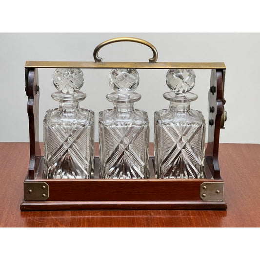 Three cut crystal decanters with decorative stoppers are displayed in the Antique English Edwardian Betjemann’s Cut Crystal Three Bottle Tantalus, c. Early 20th Century by Pearl City Antiques, set atop a wooden surface.