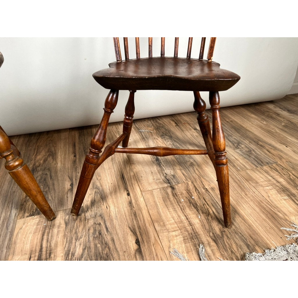 A close-up of an Antique Near Pair 18th Century Windsor Chair shows a spindle back, curved legs, and dark brown finish on a wood-patterned floor, with visible wear and part of another matching chair at the left edge. Brand: None and/or Unknown.