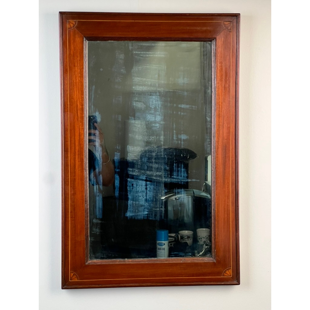 An American Federal Mahogany Wall Mirror with fan inlay and original hand-silvered glass, circa 1810–1825, reflects part of a bathroom, some toiletries, and a person holding a phone—its cloudy, streaked surface and antique craftsmanship evoke nostalgic charm reminiscent of early nineteenth-century American mirrors.