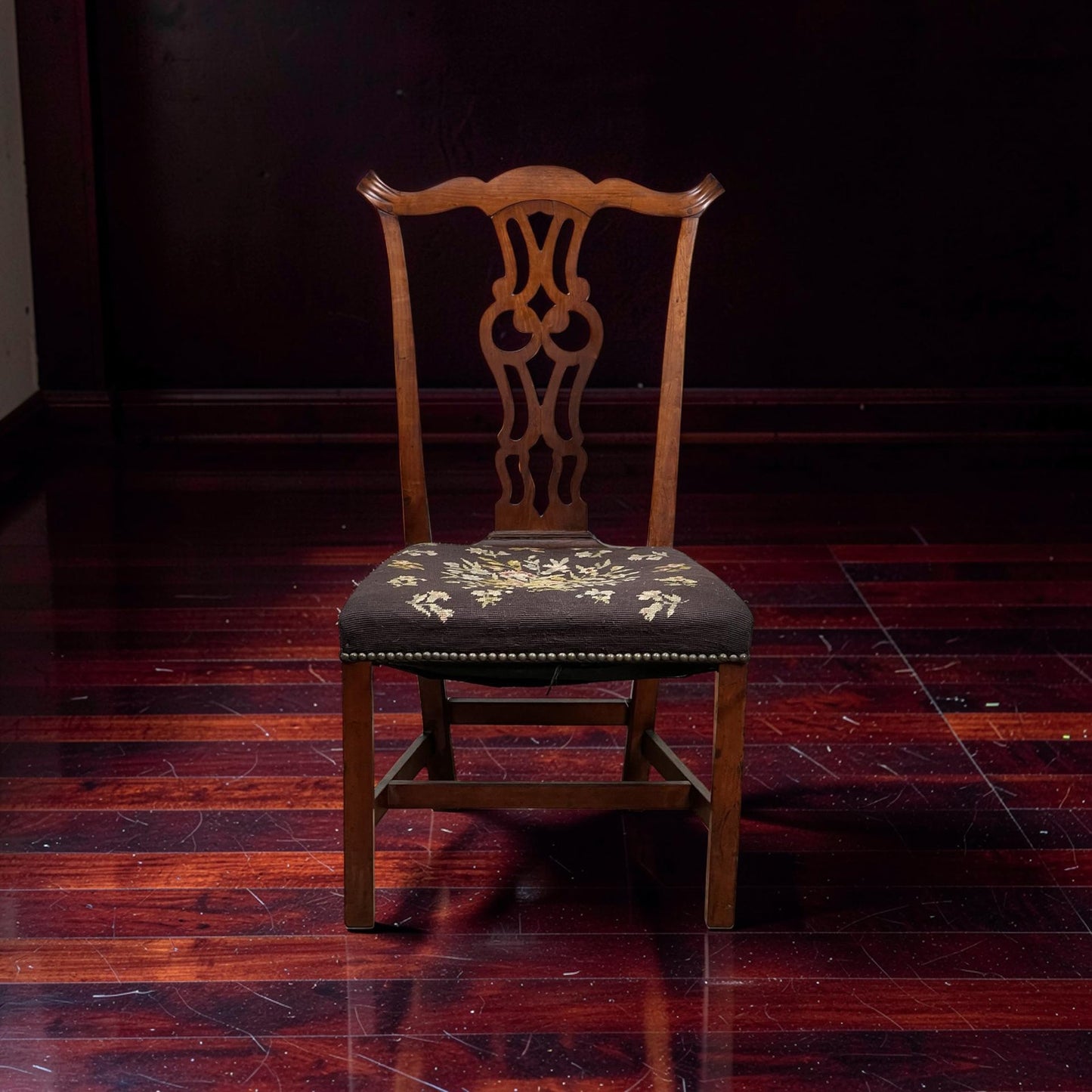 A mid-19th century Antique Chippendale Side Chair with a decorative splat back and needlepoint seat, set on a polished dark wood floor against a dark background. Brand: None/Unknown.