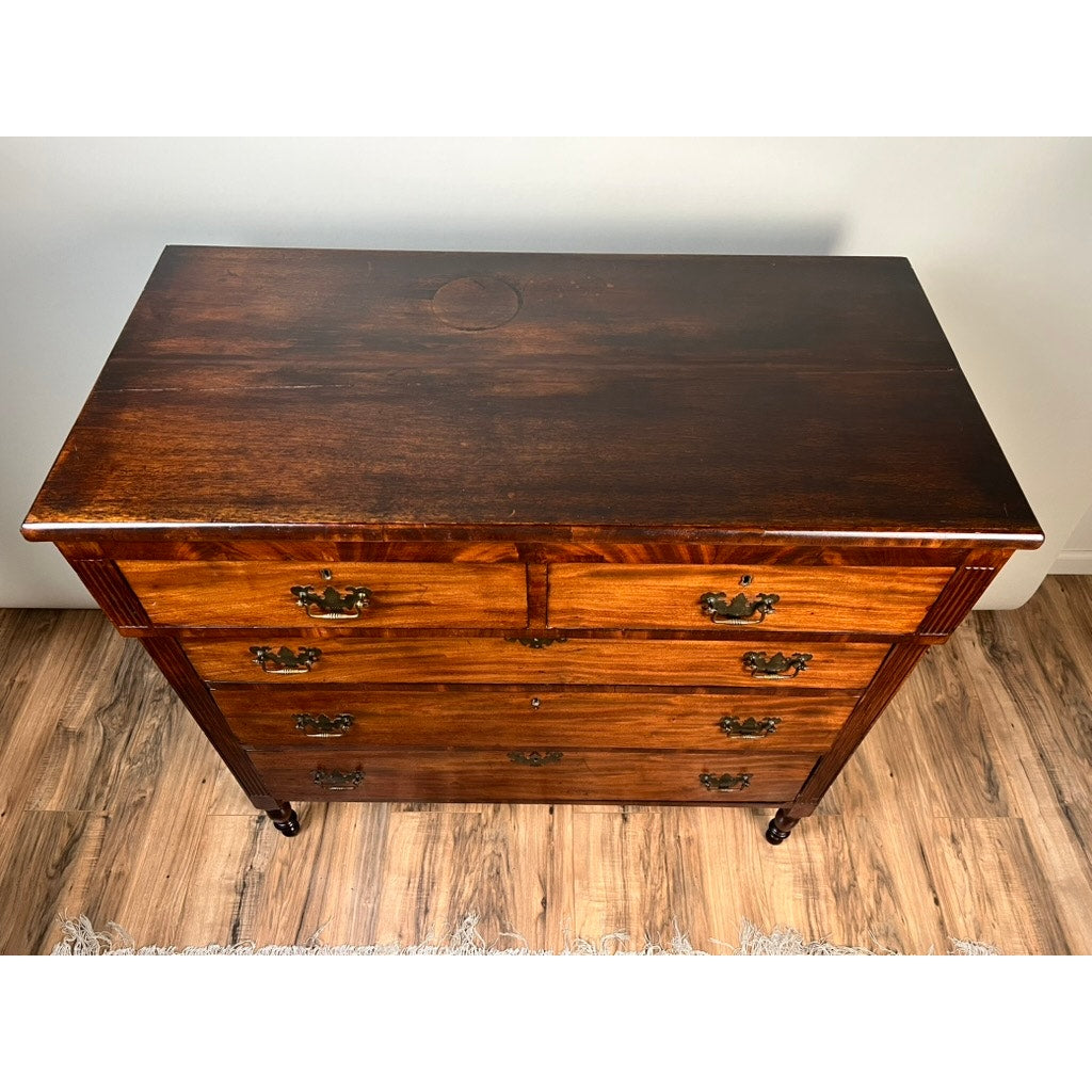 An Antique George III Sheraton Flame Mahogany Chest of Drawers (early 19th century) with four drawers, ornate brass handles, and small wheels stands on a hardwood floor beside a rug. Brand: None/Unknown.