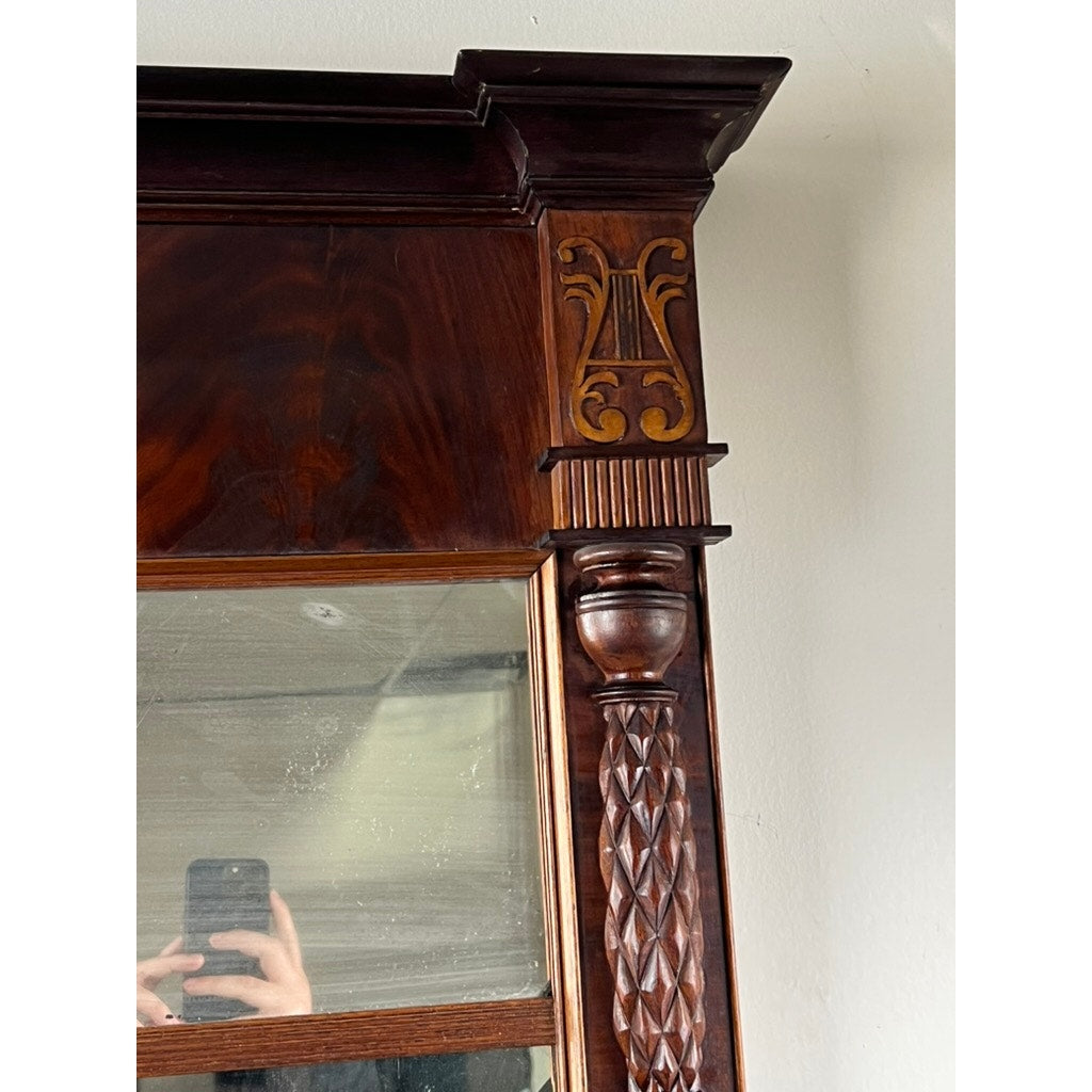 Close-up of an Antique American Empire Mahogany Pier Mirror c. 1840, showing ornate carved details like a lyre motif and fluted column; a person's hand holding a phone appears in the reflection.