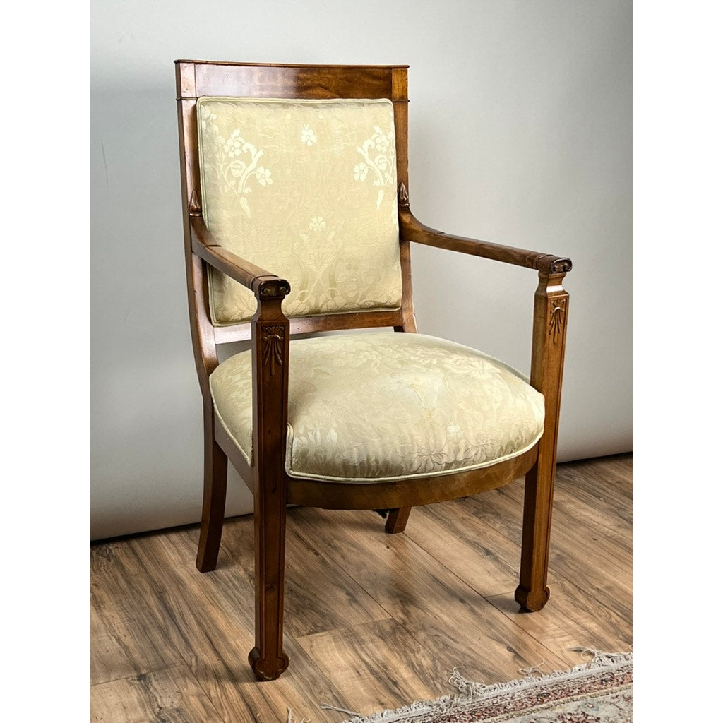 An Antique French Directoire Style Fauteuil open arm chair in Sapelle mahogany (late 19th century) with light beige, floral-patterned upholstery sits on a wood floor near a pale wall and the edge of a patterned rug.