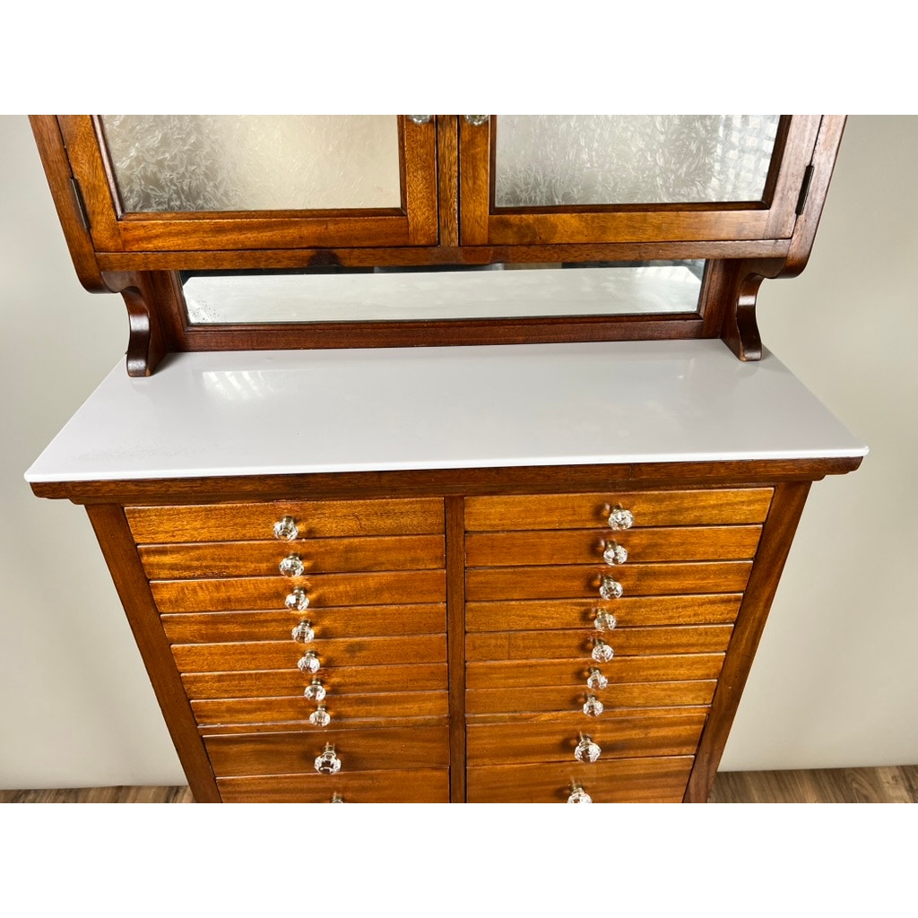 An Antique Mahogany Multi-drawer Dental Cabinet by The American Cabinet Co. (early 20th century), with glass knobs, a white countertop, and a glass-fronted upper section, stands against a beige wall.
