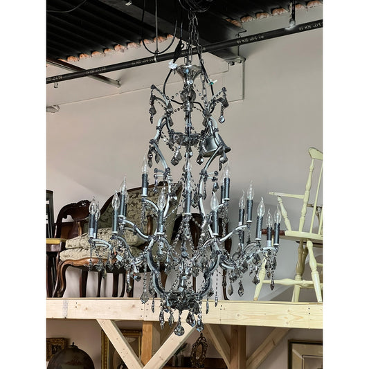 A Large Vintage Smoke Crystal 24 Light Chandelier by Pearl City Antiques hangs from the ceiling in a storage area, surrounded by vintage chairs and furniture stacked on wooden shelves.