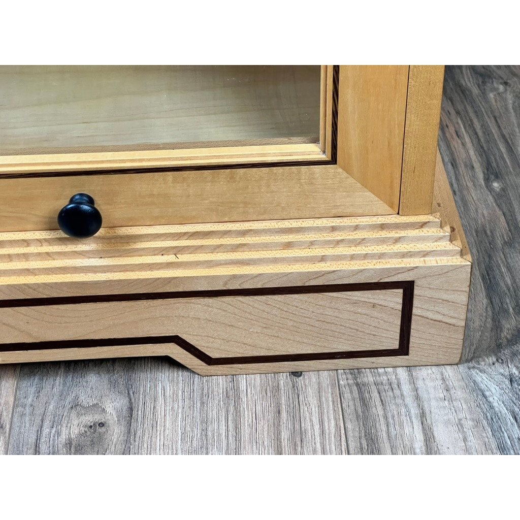 Close-up of a Vintage Bench Made Maple with Wenge Inlay Three Stack Barrister Bookcase corner, featuring a glass panel, black knob, decorative wenge inlay, and grooved maple design on a wood-textured floor. Brand unknown.