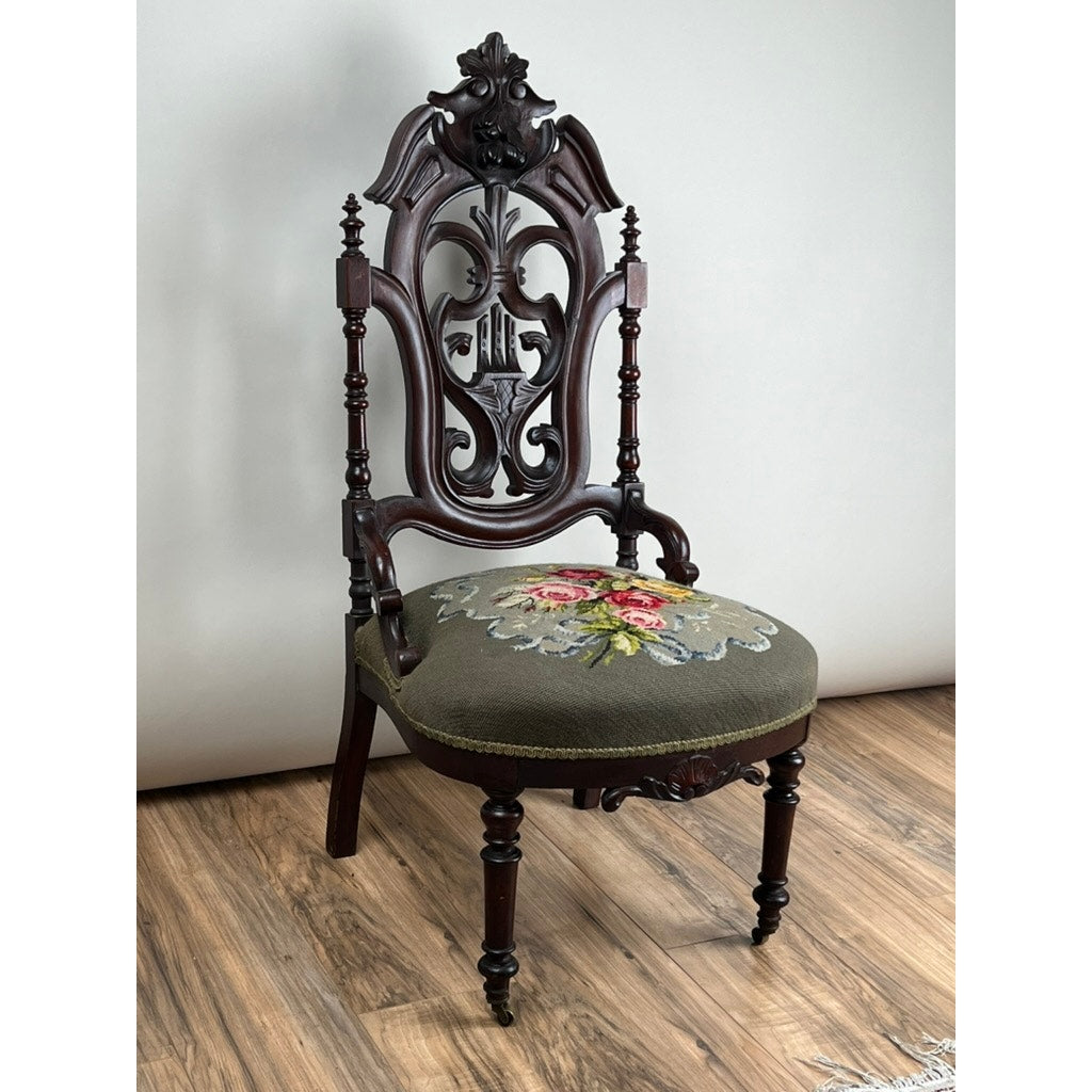 An Antique Victorian Rococo Revival Carved Walnut Slipper Chair with a floral needlepoint seat (late 19th c.) is displayed on a wooden floor against a plain light background. Brand: None and/or Unknown.