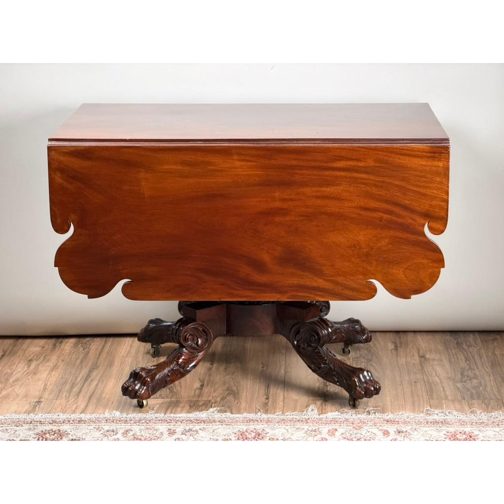 American Classical Carved Mahogany Drop-Leaf Breakfast Table, c. 1825–1840, featuring a mahogany finish, pedestal base, and carved feet with casters.