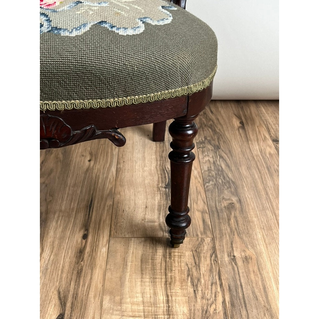A close-up of an Antique Victorian Rococo Revival Carved Walnut Slipper Chair (late 19th c.) shows the leg on a wooden floor, green upholstery with decorative trim, and a floral needlepoint seat. Brand: None/Unknown.
