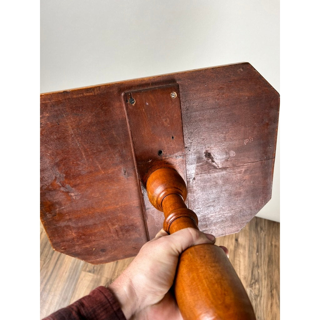 A hand holds the underside of an Antique 19th Century Cut Corner Hepplewhite Candle Stand with a square, polished base and turned handle, shown above a wood floor and in front of a white wall.