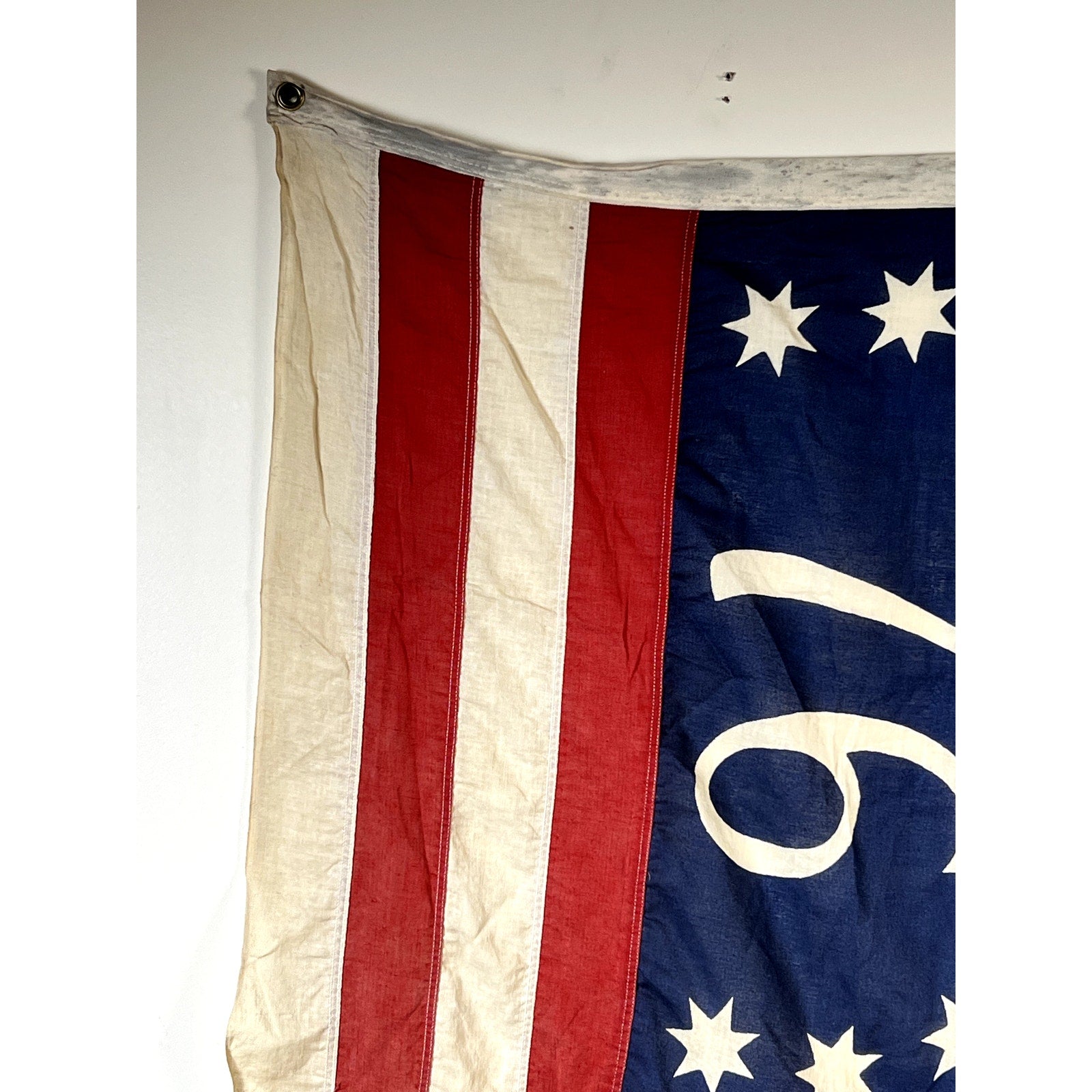 Partial view of a Vintage Valley Forge “76” Bennington American Flag from the Bicentennial Era, c. 1976, featuring red and white stripes on the left and a blue field with white stars and the number “76” on the right, hanging on a white wall.