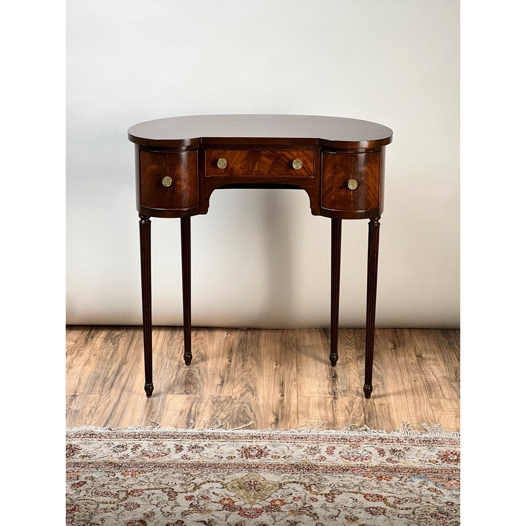 A Vintage Edwardian Sheraton Revival kidney-form dressing table c. 1940, featuring mahogany veneers, three drawers with brass knobs, tapered legs, and a polished finish, sits on a wooden floor beside a patterned rug and plain light background.