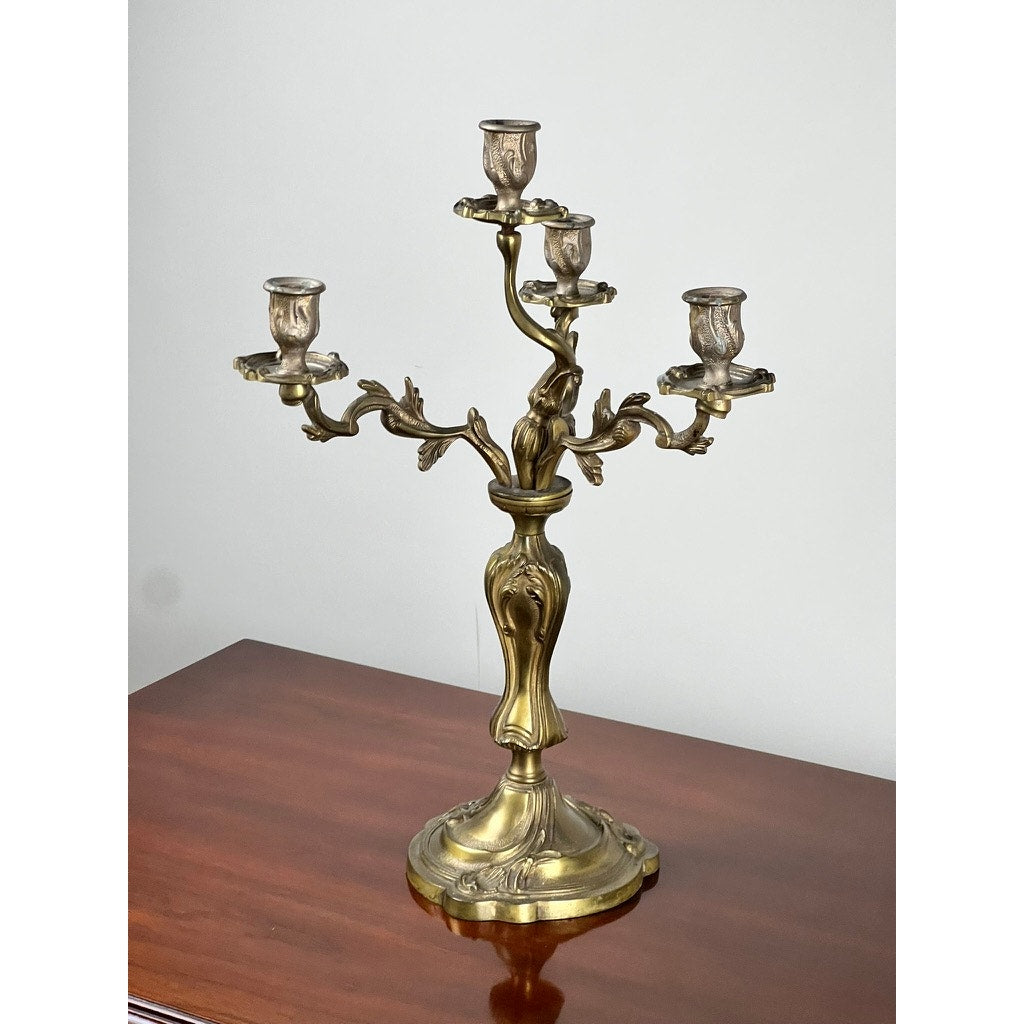 A vintage brass Louis XV style four-light candelabra with ornate, curved arms rests on a polished wooden surface against a plain light-colored wall; brand is unknown.