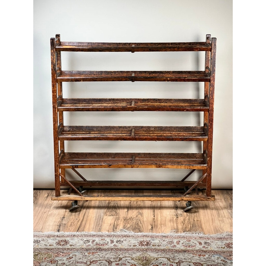 An Antique Oak Shoe Factory Rolling Rack on original industrial casters (late 19th century, brand unknown) with five angled shelves stands on a wood floor before a light wall; part of a patterned rug appears at the bottom.