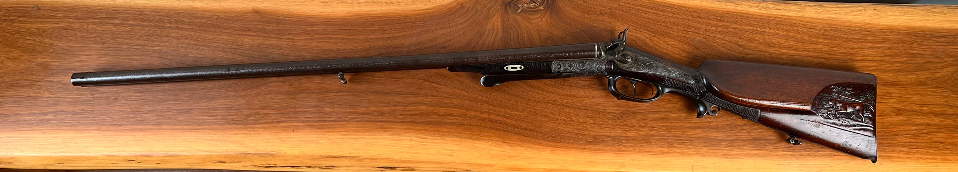 An Antique Christoph Funke Suhl German Damascus Double Barrel Hammer Shotgun, c. 1880, from Pearl City Antiques, showcases intricately carved details on its wooden stock and elegant Damascus barrels, beautifully displayed on a polished wooden surface.