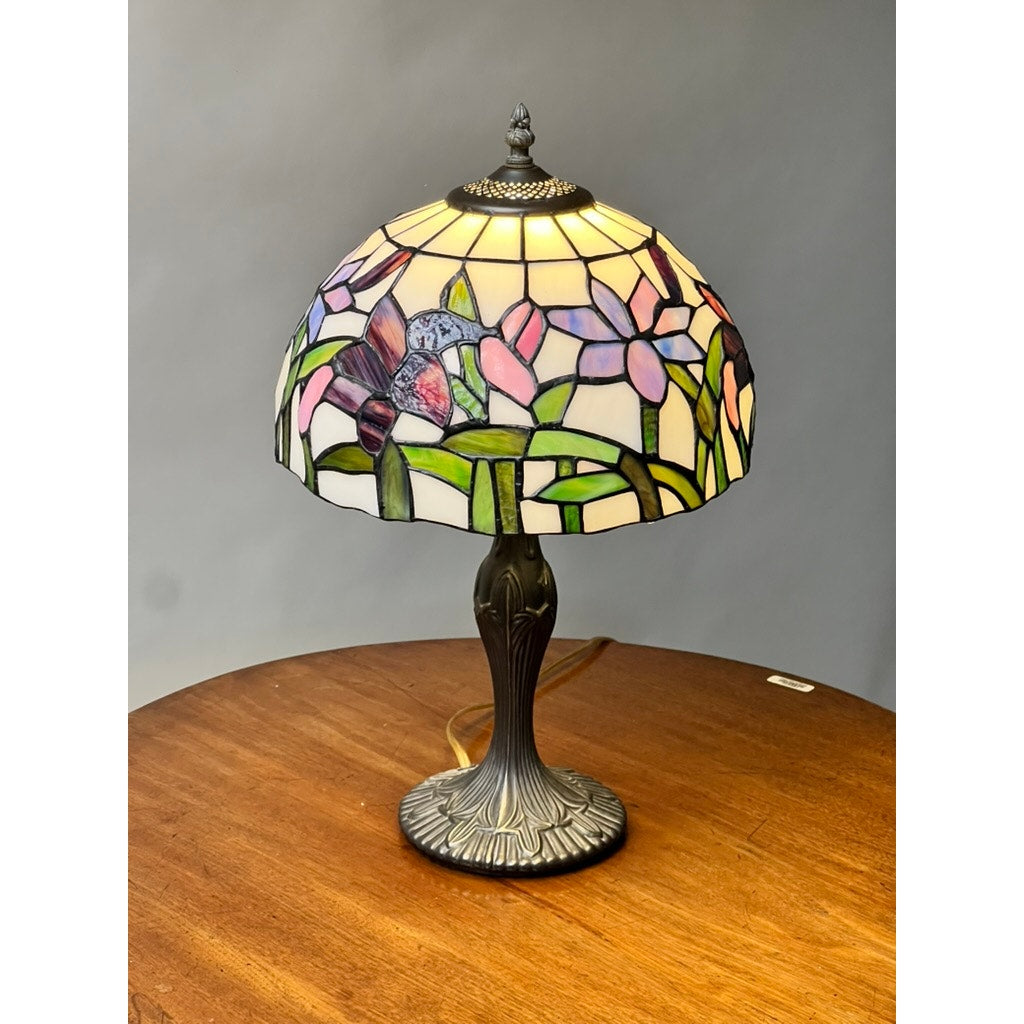 A Vintage Tiffany Style Floral Stained Glass Table Lamp with a dome featuring hummingbirds sits on a round wooden table against a plain gray background. Brand name is unknown.