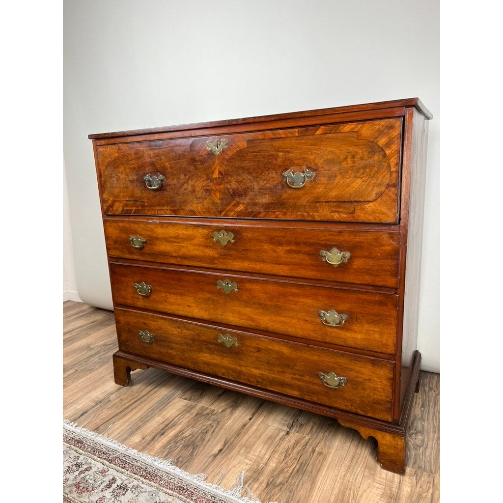An Antique George IV Butlers Secretary from the early 19th century with polished wood, four drawers, ornate brass handles, and bracket feet stands on a hardwood floor next to a patterned rug and light-colored wall. Brand: Unknown.