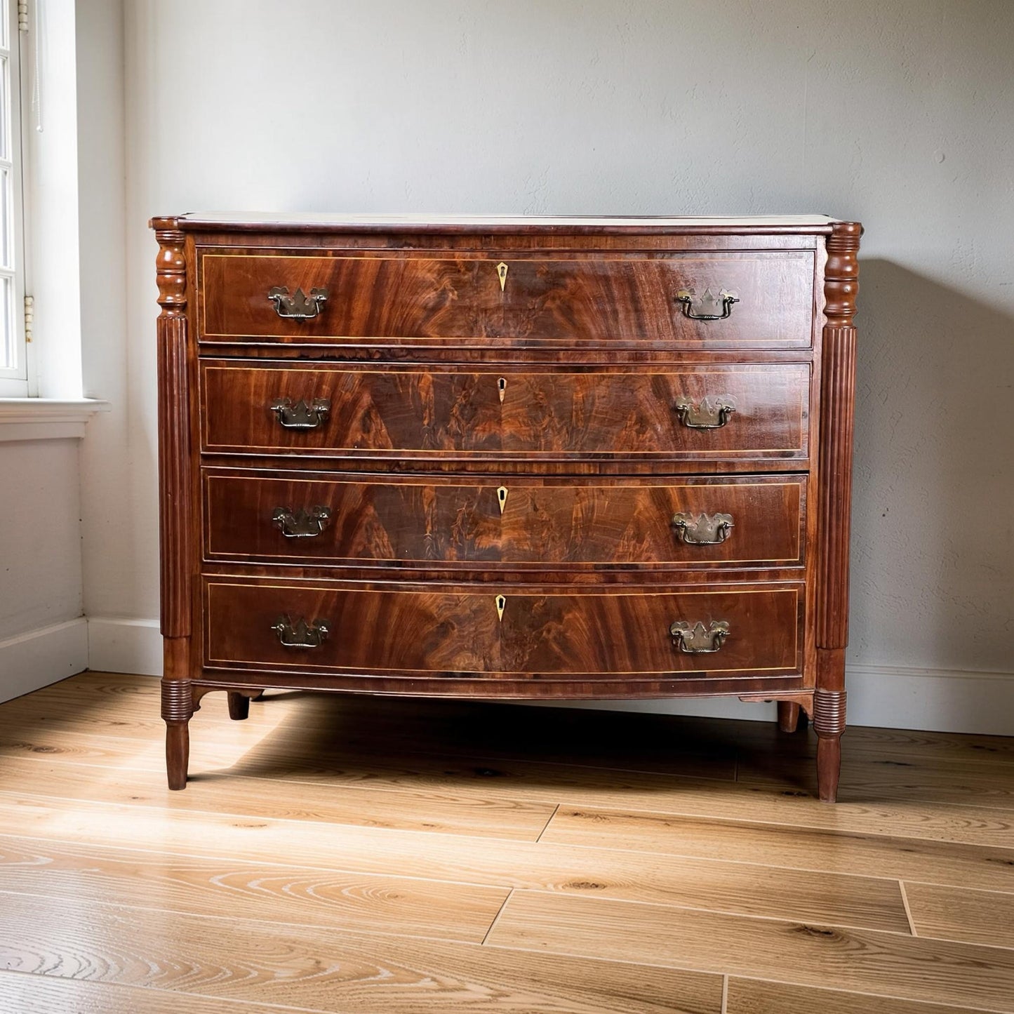 An Antique Sheraton Mahogany Bowfront Chest from the early 19th century features four drawers with brass handles and elegant wood grain, standing on slender legs in a room with light wood floors and a nearby window.