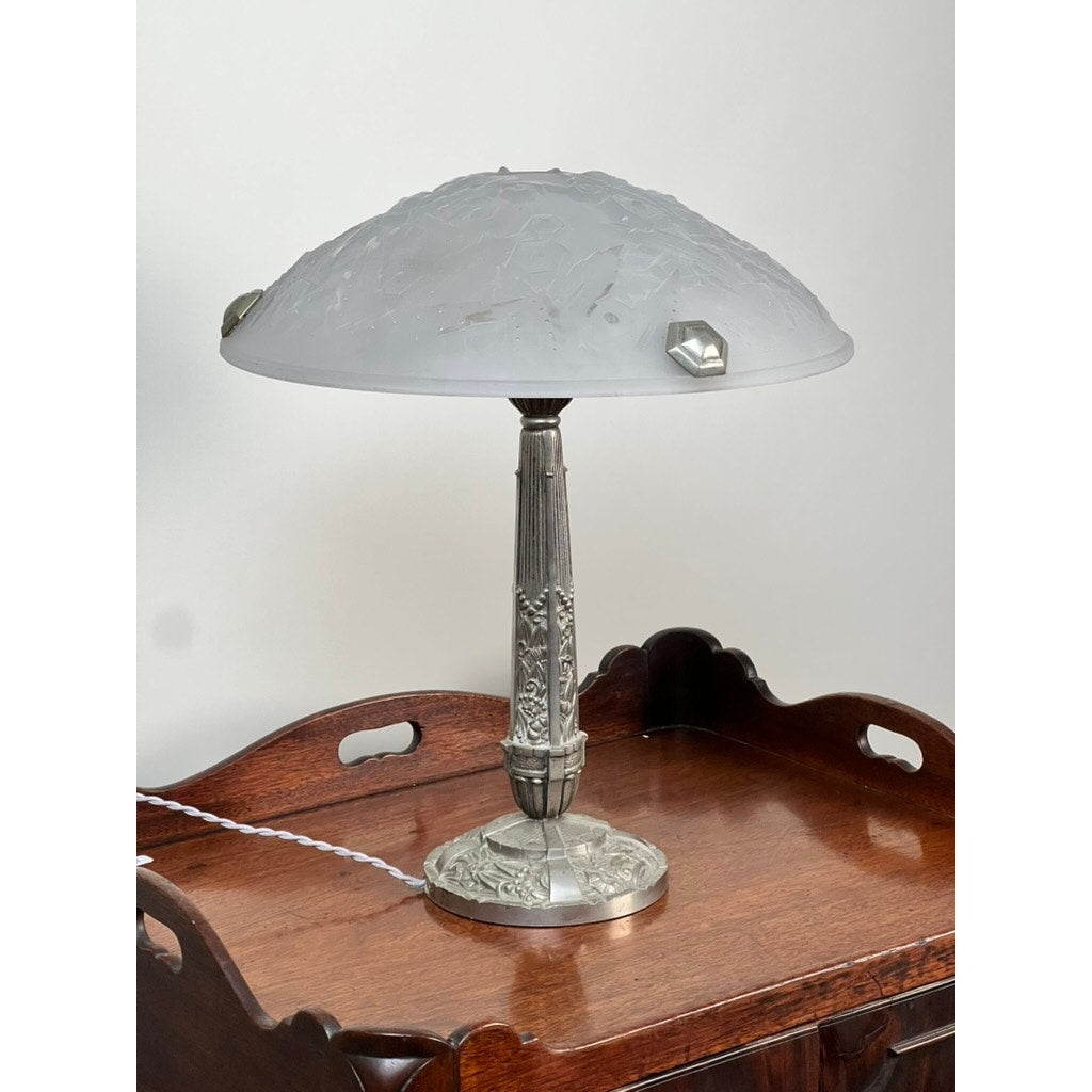 A Vintage French Art Deco Frosted Glass Table Lamp by Muller Frères Lunéville c. 1920 with an ornate silver base sits on a polished wooden tray table, plugged in with a white cord. Brand is unknown.