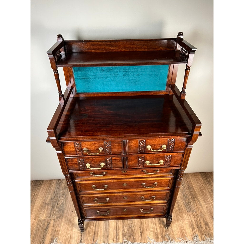 An Antique Victorian Aesthetic Movement Mahogany Four Over Four Gentlemen’s Chest c. 1880, featuring brass handles and a two-tiered upper shelf with turquoise fabric backing, sits on a wooden floor against a plain wall.