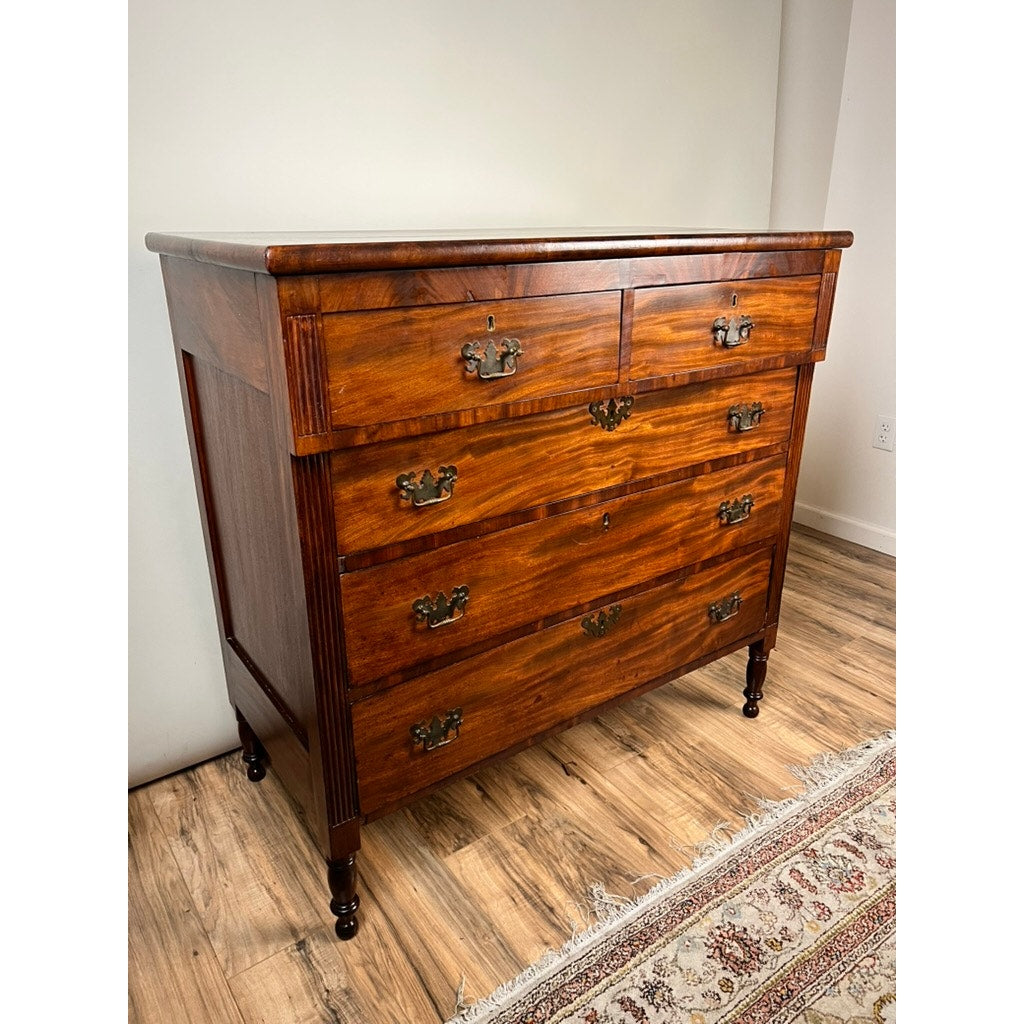 An Antique George III Sheraton Flame Mahogany Chest of Drawers from the early 19th century, featuring five drawers with brass handles, stands on turned legs over a hardwood floor next to a patterned rug in a well-lit room.