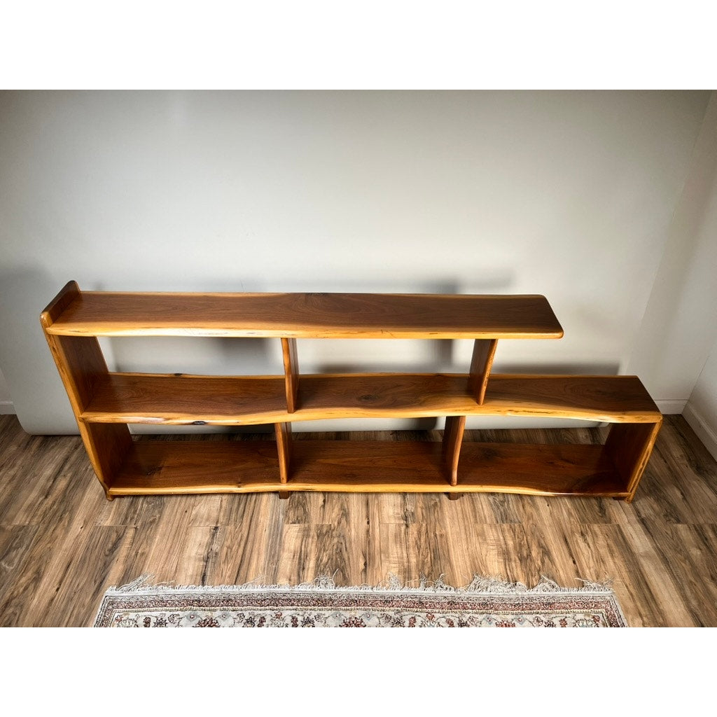 The Vintage Mid-Century Modern Style Live Edge Walnut Bookshelf features three open shelves of varying heights and lengths, positioned against a white wall on a wooden floor with part of a patterned rug in the foreground.