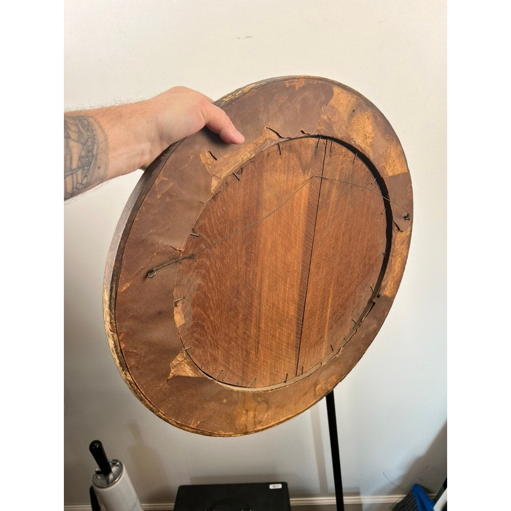 A hand holds the back of an antique oval “Storm Tossed Ship” oil on board painting, late 19th century, showing worn wood, cracks, tattered brown paper, and old wiring—evidence of its age and storied maritime past.