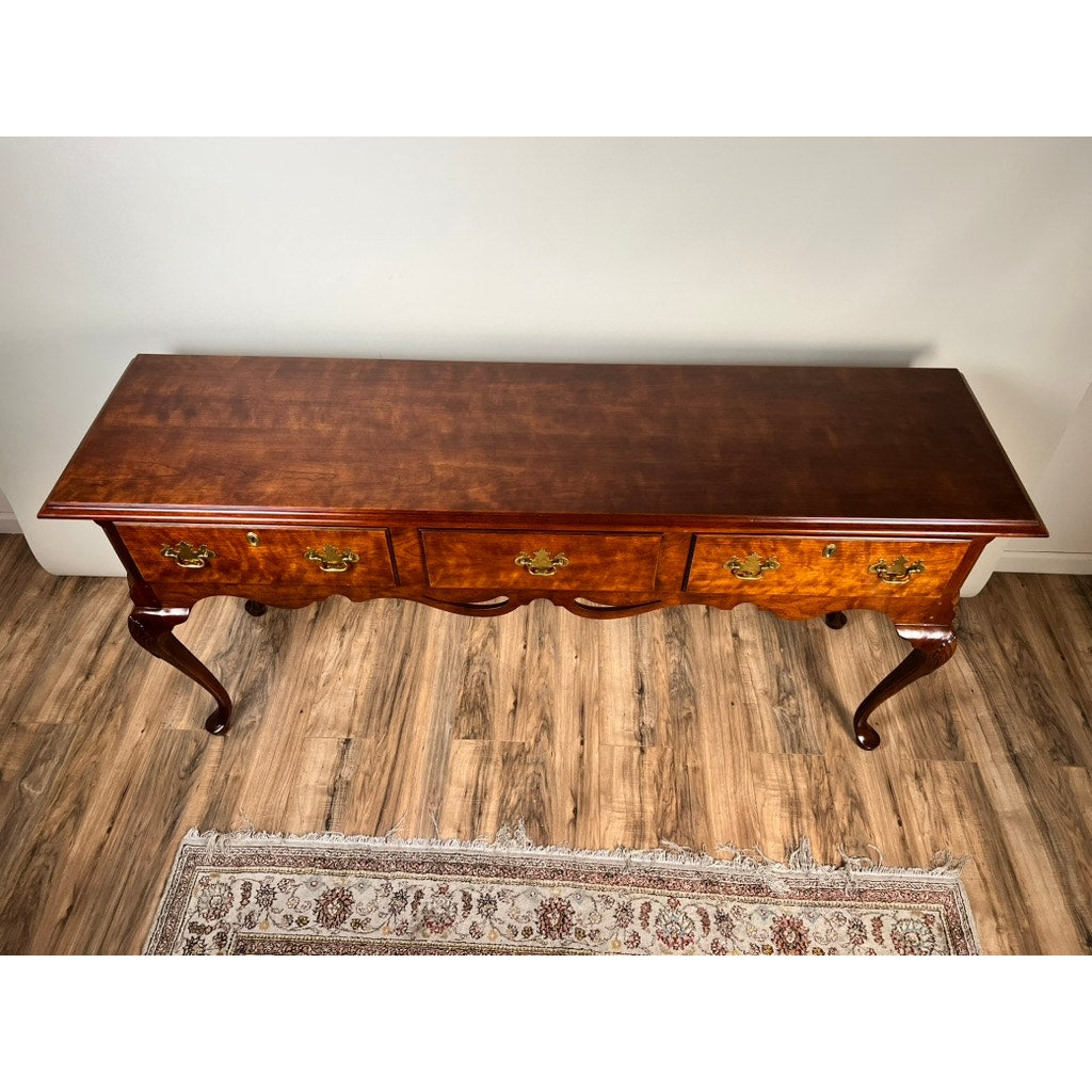 A Vintage Solid Cherry Queen Anne Style Sideboard by Henredon, featuring three drawers and curved legs, stands on a wooden floor beside a patterned rug and against a plain white wall.