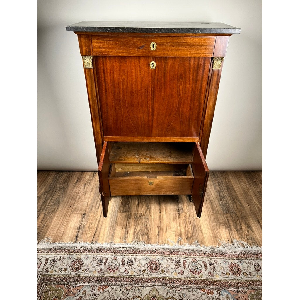 An Antique French Empire Secrétaire Abattant with ormolu mounts, mahogany veneer, dark stone top, and two open lower shelves stands on a wooden floor beside a patterned area rug. Mid 19th century; maker unknown.