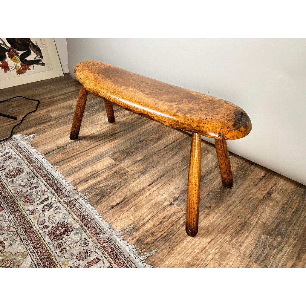 The Vintage Hand Made Oak Wabi Sabi Bench, signed by Neil Panyik, features a smooth oval seat and four angled legs, shown on a wood floor beside a patterned rug in a bright room.