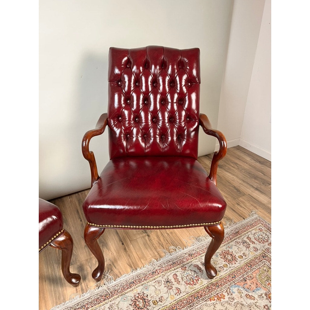 A pair of Chippendale style tufted burgundy leather library armchairs, featuring button-tufted backrests with wooden arms and legs, is shown set on a light wood floor beside a patterned rug against a plain white wall.
