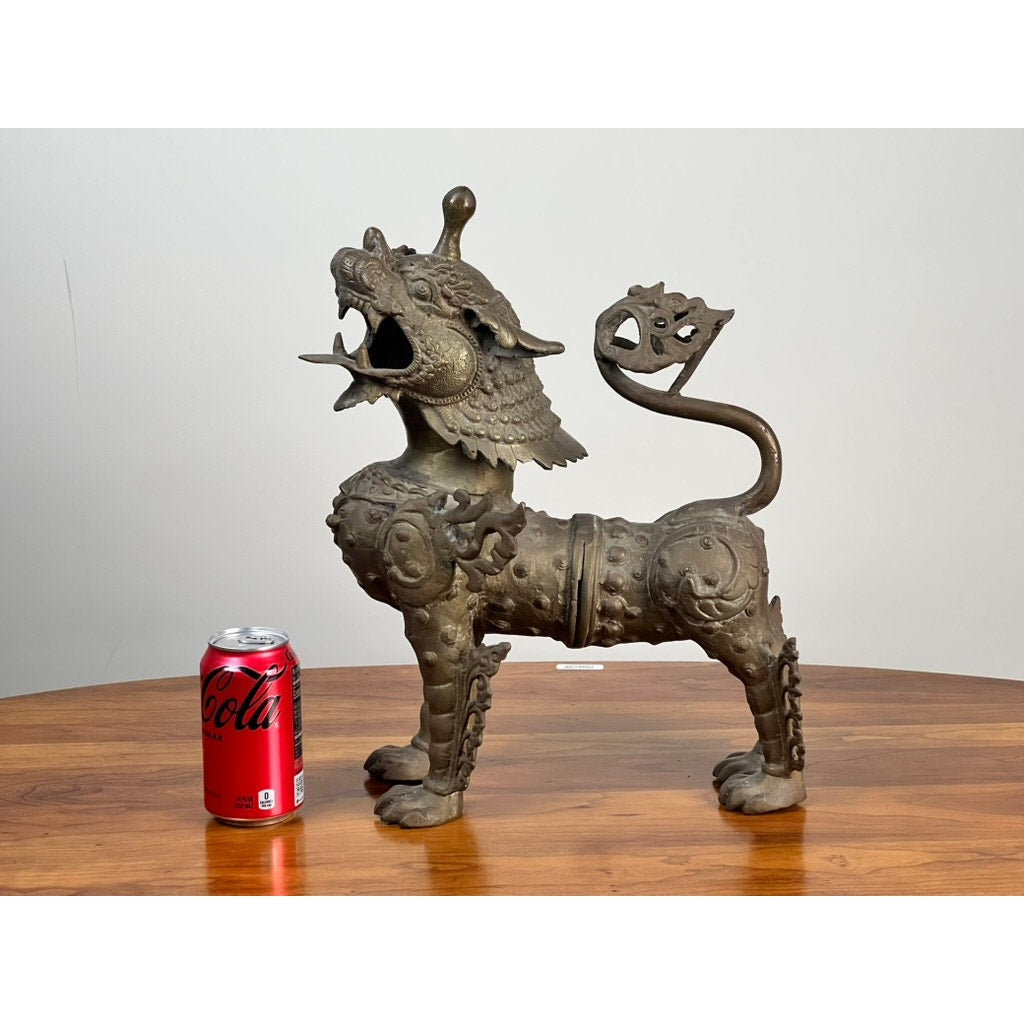 A Vintage 17” Cast Brass Pixiu Tibetan Lion c. 1960 stands on a wooden table next to a red Coca-Cola can, showcasing its size. Ideal for lovers of Tibetan Chinese decor or Asian art, it’s photographed against a plain, light background. Brand: Unknown.