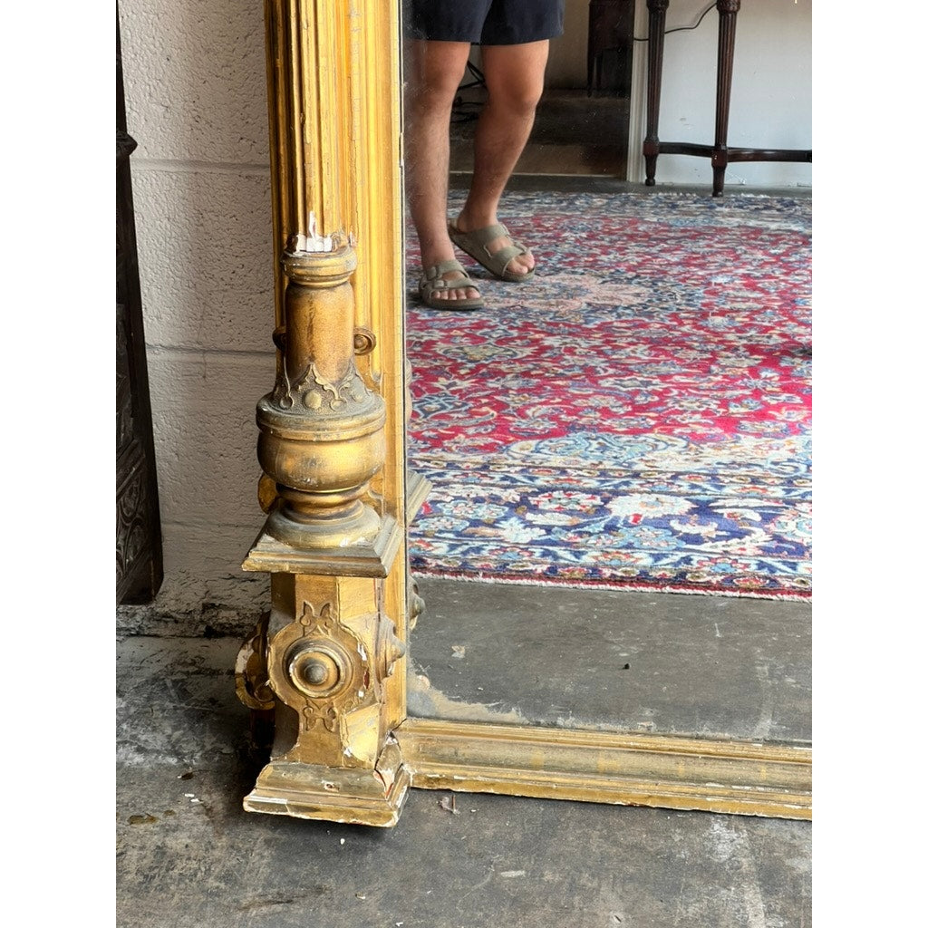 An Exceptional Monumental Victorian Renaissance Revival Pier Mirror, circa 1870–1885, reflects a person's lower legs clad in shorts and sandals as they stand on a vibrant patterned rug atop a concrete floor.