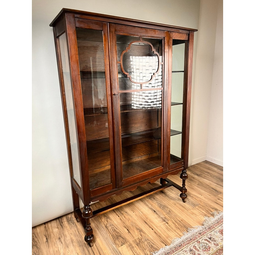 A Depression Era Mahogany Curio Display Cabinet with Glass Sides, Circa 1930s, this vintage American furniture piece features elegant glass doors and side panels, standing gracefully on four turned legs. Inside, shelves provide ample display space, while a decorative pattern accents the upper glass door. Reminiscent of classic 1930s design, it is beautifully presented on a wood floor.