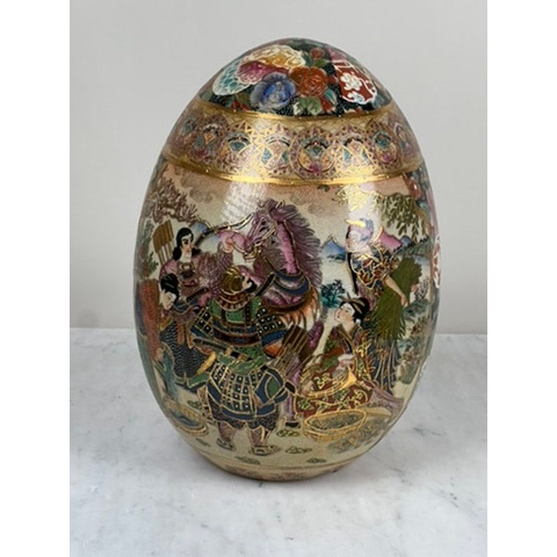 A large antique Satsuma porcelain egg, Chinese export, displays intricate samurai warrior and landscape scenes with colorful patterns and gold accents on a white background, showcased on a marble surface.