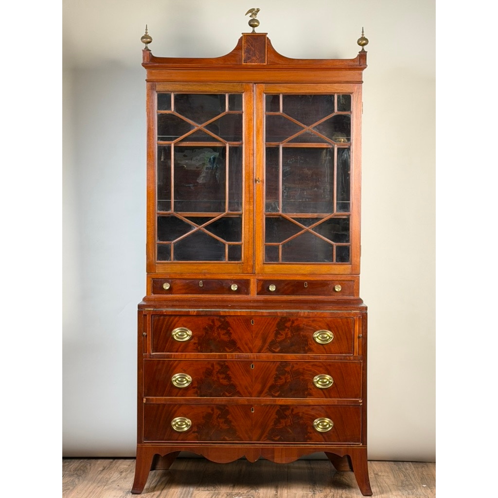 Federal Flame Mahogany Secretary Bookcase with Eagle Finial, c. 1810–1825. This early 19th-century American piece is crafted in the Federal period style, featuring glass-paneled upper doors, exquisite flame mahogany veneers, and four lower drawers accented by brass handles. The bookcase stands on short legs and is topped with a distinctive eagle finial for decorative appeal. Brand: Unknown.