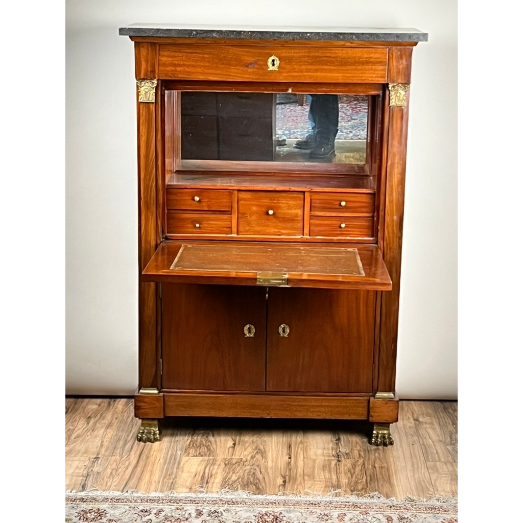 An Antique French Empire Secrétaire Abattant with ormolu mounts, c. mid 19th century, featuring mahogany veneer, marble top, mirrored back, drawers, and fold-down writing surface stands on a wood floor near a rug.