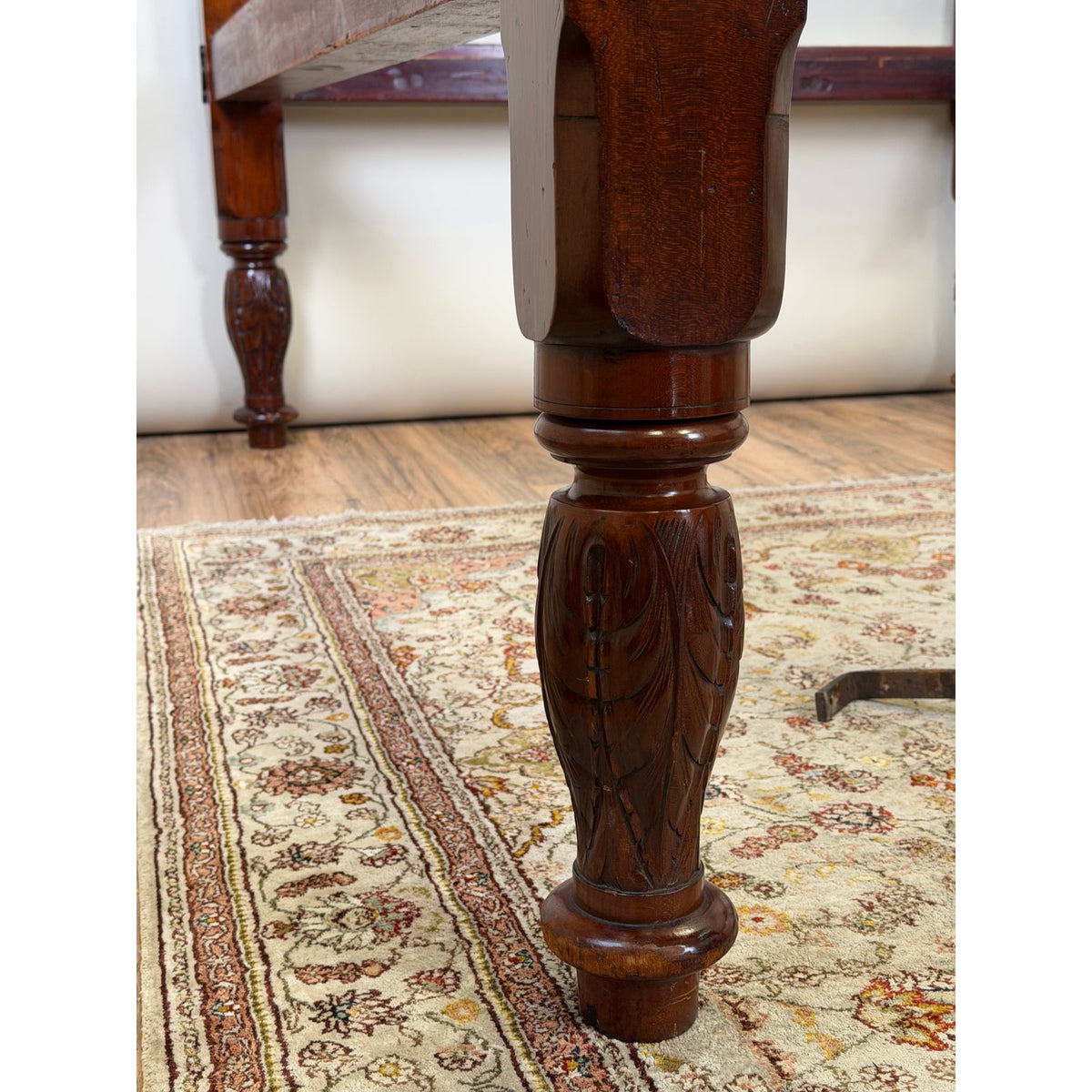 A close-up of a dark, ornate mahogany leg on an American Classical Carved Mahogany Four-Poster Tester Bed, Possibly New York, circa 1825–1840, featuring carved leaf motifs and standing on a patterned area rug.