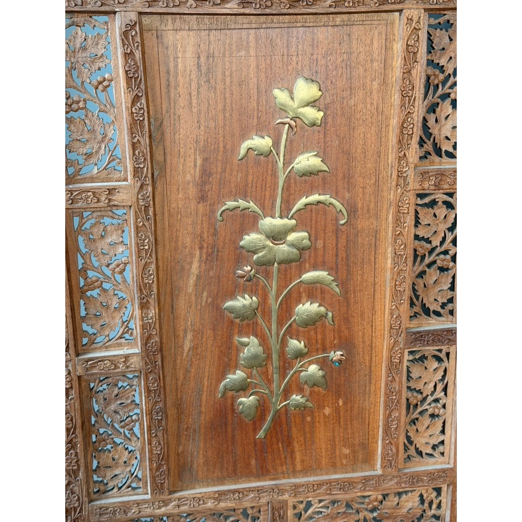 The Early 20th Century Anglo-Indian Carved Teak Four-Panel Screen from Pearl City Antiques features applied brass floral decorations at its center, complemented by intricate leaf carvings on a dark wood background.