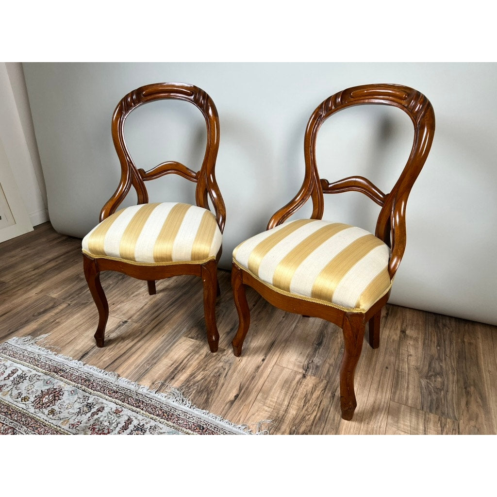 A pair of Antique Victorian Balloon Back Chairs from the late 19th century, featuring curved backs and light yellow and white striped upholstered seats, are set on a wooden floor beside a patterned rug.