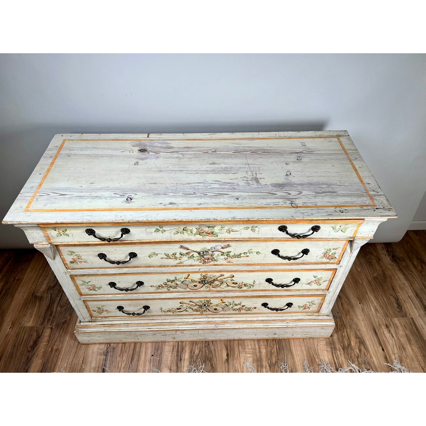 An Antique Italian Paint Decorated Four Drawer Commode from the early 19th century, featuring ornate hand-painted designs and black metal handles, stands on a wooden floor against a plain white wall. Brand unknown.