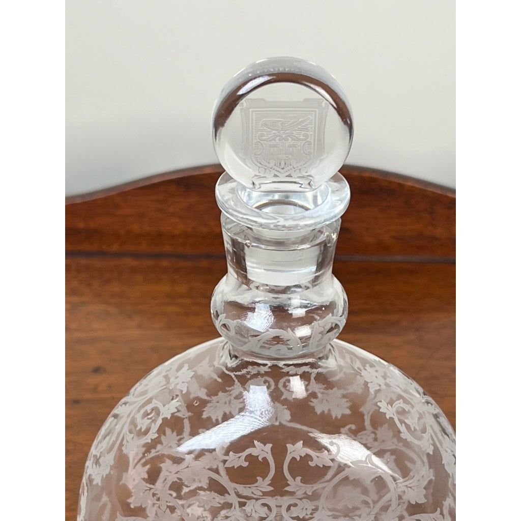 A close-up of a Vintage Baccarat "Michelangelo" Crystal Decanter for Martell Cognac c. 1930, featuring intricate floral etching and a round stopper engraved with a heraldic shield, set against a wooden background.