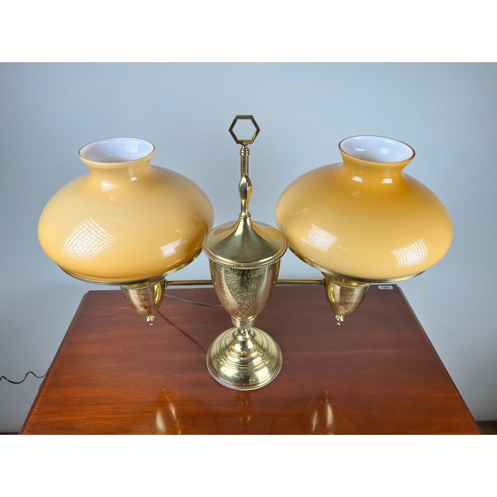 An Antique Bradley and Hubbard Brass Double-arm Student Lamp with carmel shades by Bradley and Hubbard is displayed on a polished wooden surface against a light-colored wall.