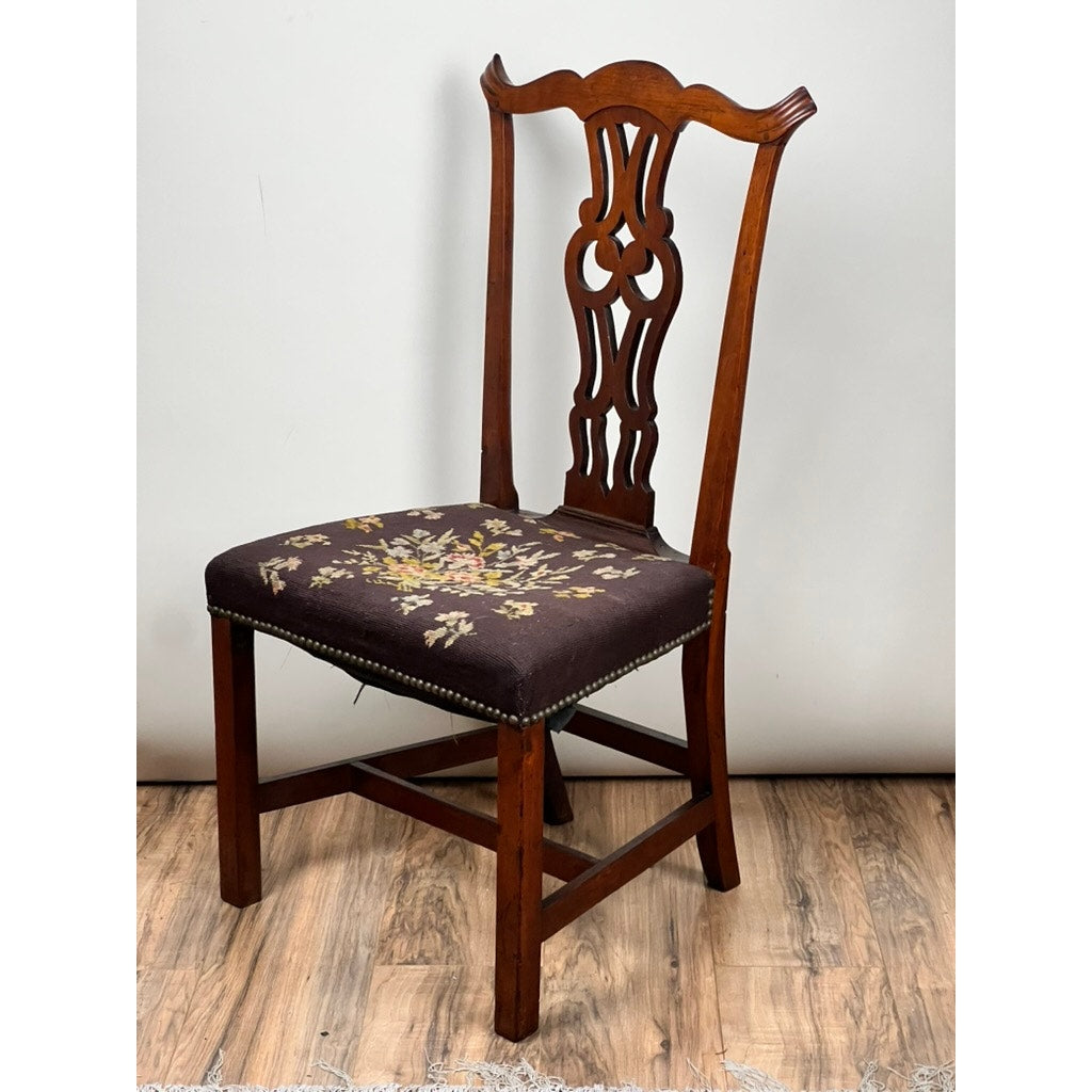 A mid-19th century Antique Chippendale Side Chair with a carved backrest and floral needlepoint upholstered seat, displayed on a wooden floor against a plain light-colored wall. Brand: None/Unknown.