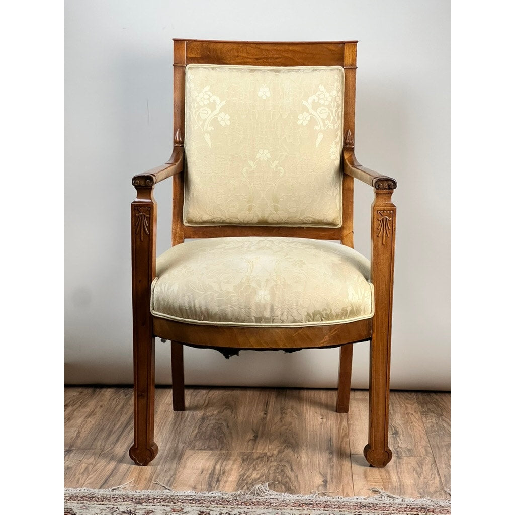 An Antique French Directoire Style fauteuil open arm chair in sapelle mahogany (late 19th century) with carved details and cream floral upholstery sits on a wooden floor against a plain white wall.