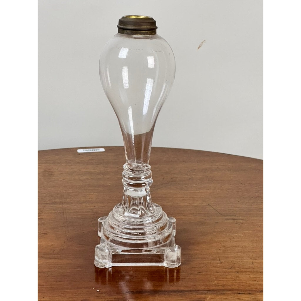 An Attributed Boston & Sandwich Pressed Glass Boudoir Fluid Lamp, circa 1850—crafted of clear glass with a brass burner and characterized by its bulbous top, slender neck, and square pedestal base—is displayed on a round wooden table. This refined piece exemplifies mid-19th century American lighting design.