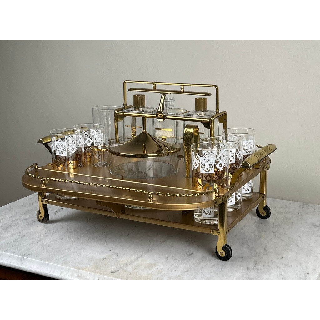 A Vintage Mid Century Modern Bar Caddy from the 1960s with wheels displays glassware—a decanter, white-patterned cocktail glasses, a lidded dish, and a gold-handled pitcher—on a marble top against a plain wall. Brand: Unknown.