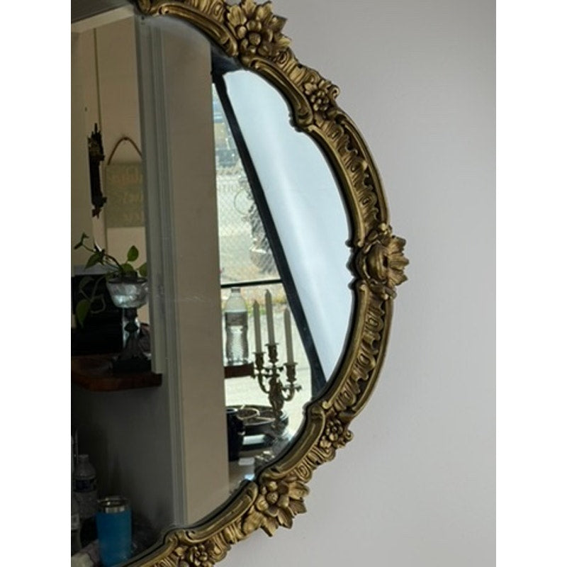 An Antique Gilt Wood Oval Mirror (c. Early 20th Century) with a gold frame reflects a candlestick holder, plant, and household items near a window; the plain white wall beside it enhances its ornate design.